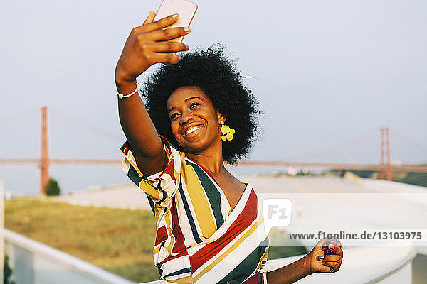 Selbstbewusste junge Frau  die ein Selfie mit ihrem Smartphone macht  mit der Brücke 25 de Abril im Hintergrund