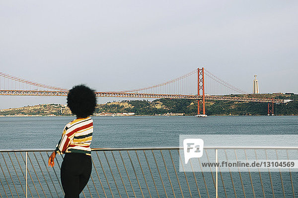 Junge Frau steht am Ufer und blickt auf die Brücke 25 de Abril und die Cristo Rei-Statue  Lissabon