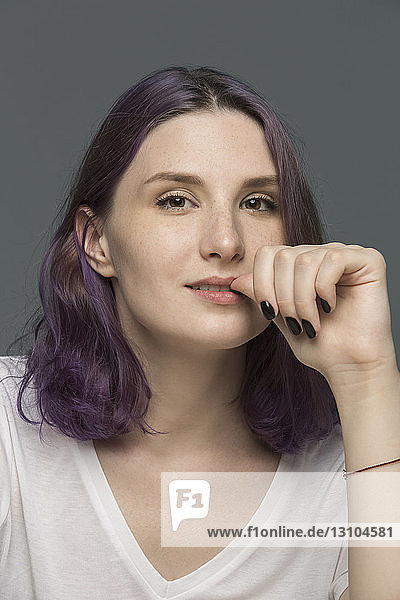 Porträt einer jungen Frau mit gefärbten Haaren beim Nägelkauen vor grauem Hintergrund