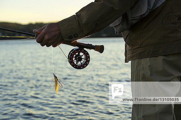 Eine Nahaufnahme der Hände eines Fliegenfischers  der seine Fliegenrute und -rolle hält  und einer Lefty's Deceiver -Streamerfliege.