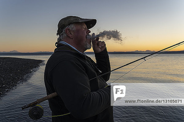 Ein älterer kaukasischer Fliegenfischer genießt eine Zigarre bei Sonnenaufgang  bevor er an der Westküste der USA mit der Fliege auf Cutthroat-Forelle und Lachs fischt