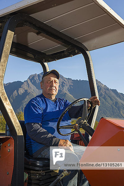 A farmer wearing a blue shirt sits on an old red tractor in an open field Pioneer Peak in the background South-central Alaska; Palmer Alaska United States of America