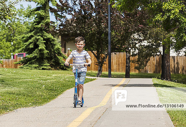 Young boy riding his scooter on a path in a city park and smiling for the camera: Edmonton  Alberta  Canada