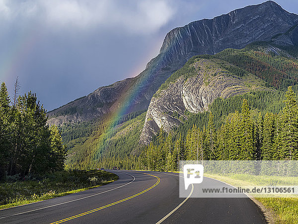 Straße durch die zerklüfteten kanadischen Rocky Mountains mit einem Regenbogen unter den Gewitterwolken; Alberta,  Kanada