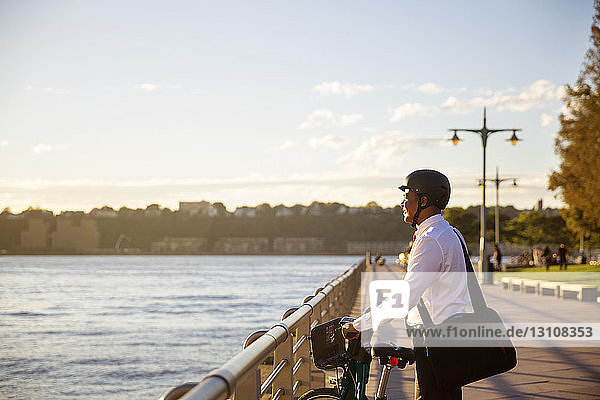 Businessman with bicycle looking away while standing by river during sunset