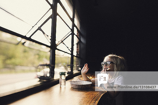 Girl wearing eyeglasses having food while sitting at table by window in darkroom
