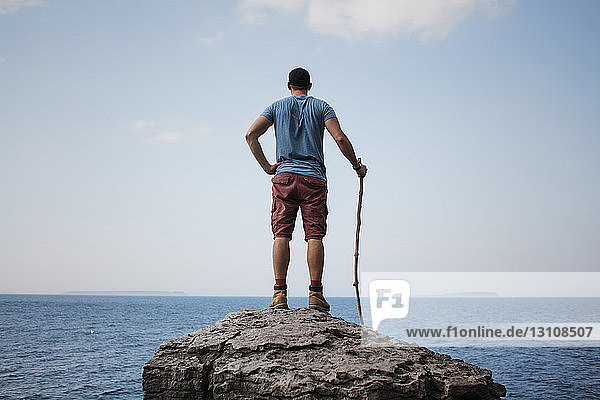 Rear view of hiker with hand on hip holding stick while standing on rock against sea and sky at Bruce Peninsula National Park