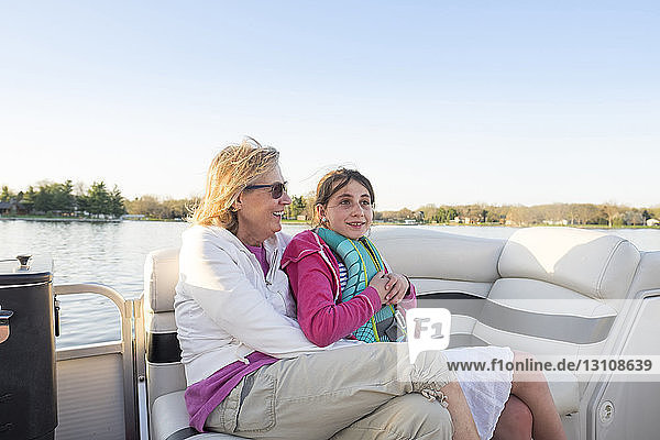 Happy grandmother with granddaughter sitting in boat on lake against clear sky