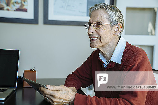 Lächelnder älterer Mann schaut weg  während er einen Tablet-Computer im Büro des Finanzberaters hält