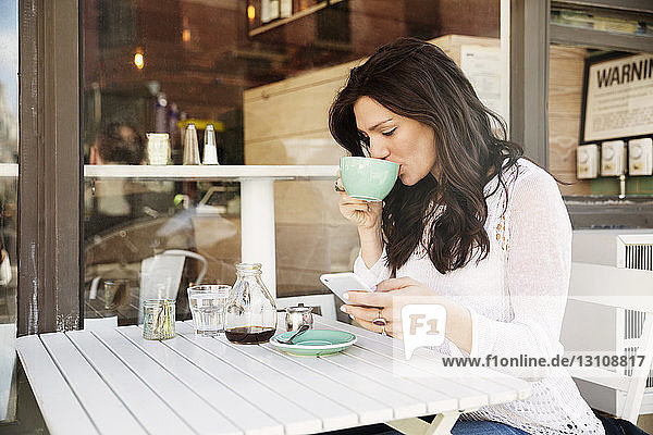 Frau trinkt Kaffee  während sie im Straßencafé ein Smartphone benutzt
