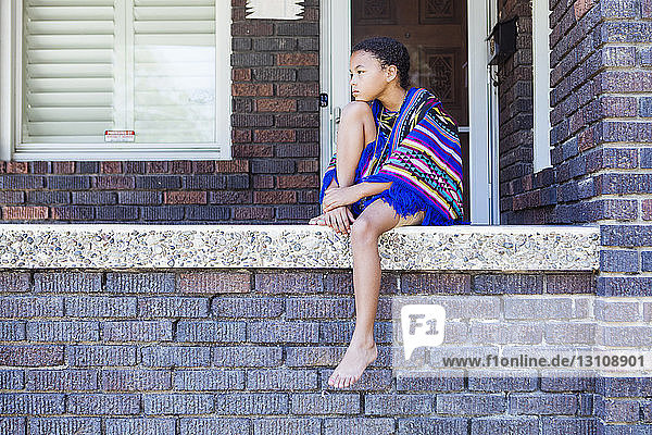 Full length of thoughtful girl with blanket looking away while sitting on retaining wall