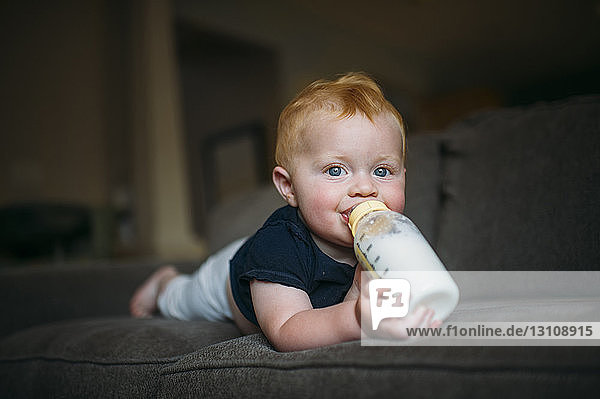 Porträt eines süßen kleinen Jungen  der Milch aus der Flasche trinkt  während er zu Hause auf dem Sofa liegt