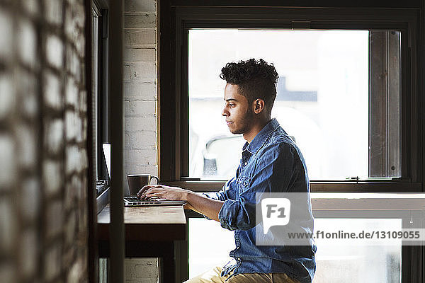 Seitenansicht eines ernsthaften Mannes  der einen Laptop benutzt  während er an den Fenstern eines Cafés sitzt