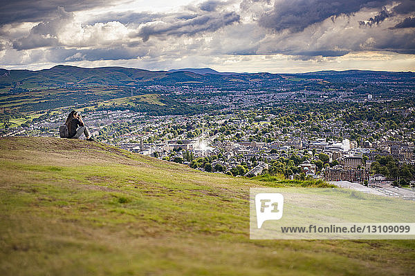 Junge Frau fotografiert  während sie auf einem Hügel am Stadtbild vor bewölktem Himmel sitzt