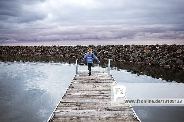 Rear view of girl walking on jetty over lake against cloudy sky