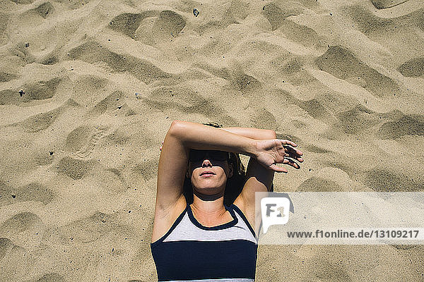 Draufsicht einer Frau  die ihr Gesicht bedeckt  während sie am sonnigen Tag auf Sand liegt
