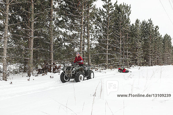 Brother towing sister with inflatable ring while driving quad bike on snow covered field