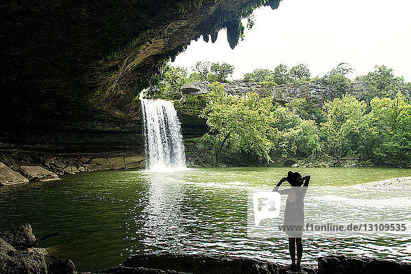 Frau betrachtet malerischen Wasserfall im Wald vor klarem Himmel