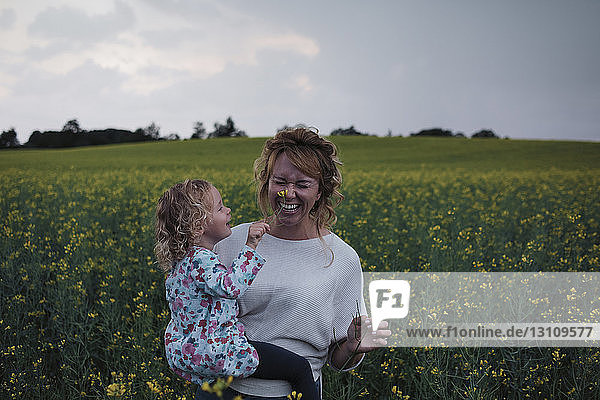 Tochter berührt Blume auf fröhlicher Nase der Mutter auf Feld gegen Himmel