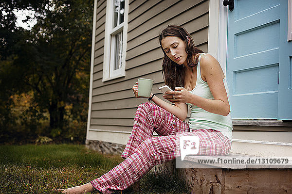 Junge Frau benutzt Smartphone  während sie vor dem Haus sitzt