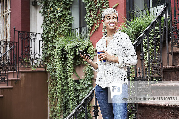 Happy woman looking away while holding mobile phone and coffee cup
