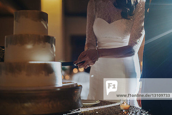 Midsection of bride cutting wedding cake while standing by bridegroom in hotel