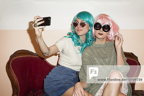 Women wearing sunglasses clicking selfie while sitting on couch against wall