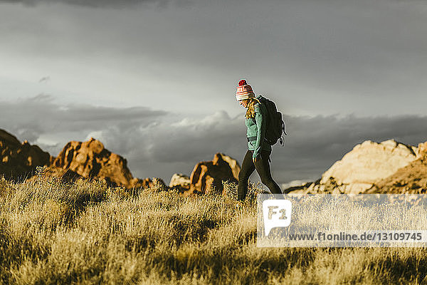 Seitenansicht einer Wanderin mit Rucksack auf einem Grasfeld vor Felsformationen und bewölktem Himmel bei Sonnenuntergang