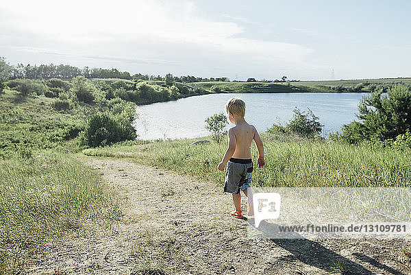 Rear view of shirtless boy walking on field by lake against sky