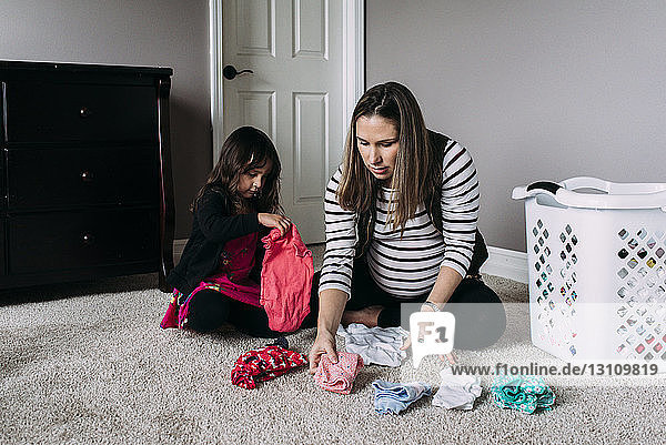 Schwangere Mutter mit Tochter faltet Kleidung  während sie zu Hause auf dem Teppich sitzt