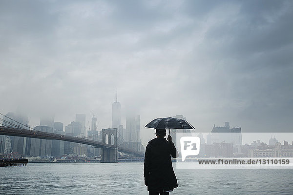 Rückansicht eines Mannes  der einen Regenschirm hält  während er die Stadtansicht vor bewölktem Himmel betrachtet