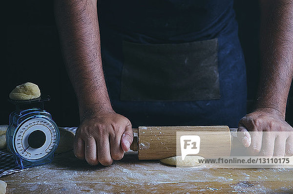 Mittelteil eines Mannes  der mit einem Nudelholz bei Tisch in einer Bäckerei Teig rollt