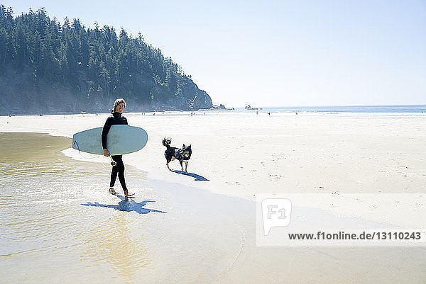 Portrait of happy man holding surfboard with dog walking at beach