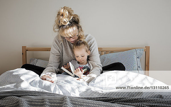 Mother with daughter reading book while sitting on bed at home