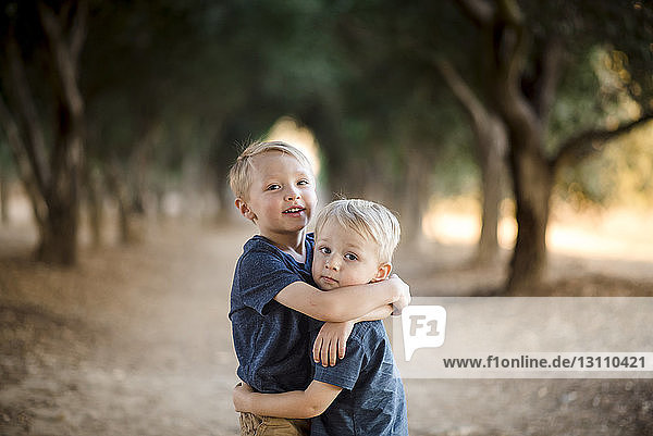 Porträt von Jungen  die sich umarmen  während sie auf einem Gehweg inmitten von Bäumen stehen