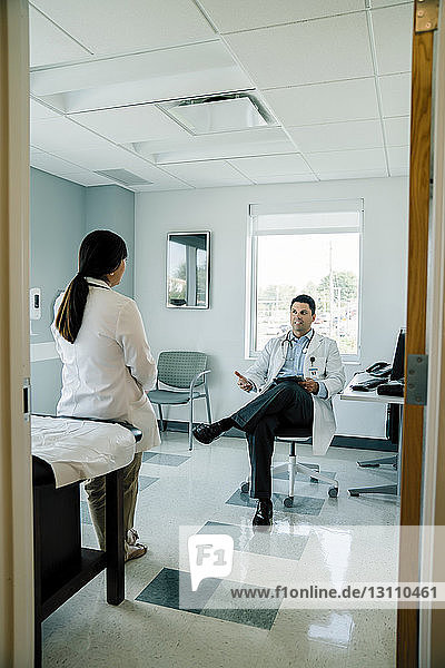 Arzt diskutiert mit weiblicher Kollegin während der Arbeit im Untersuchungsraum durch die Tür gesehen