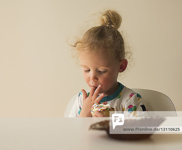 Mädchen leckt Zuckerguss von Törtchen  während sie zu Hause auf einem Stuhl an der Wand sitzt