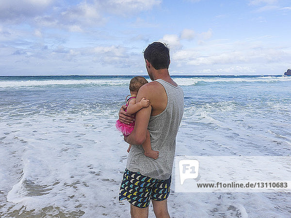 Father carrying daughter while standing at beach against sky
