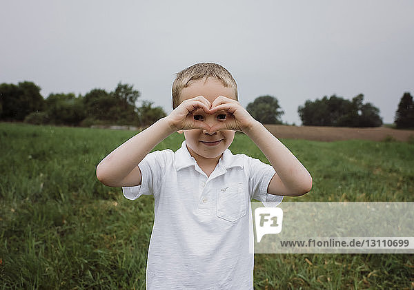 Portrait of boy making heart shape while standing on grassy field against sky at park