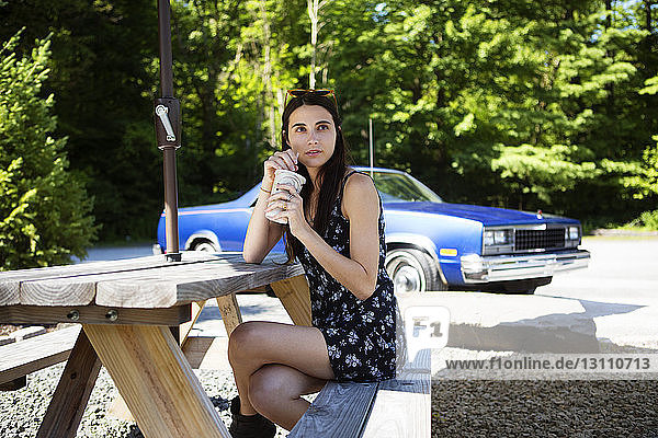 Nachdenkliche Frau trinkt Saft  während sie auf einem Picknicktisch an Bäumen sitzt