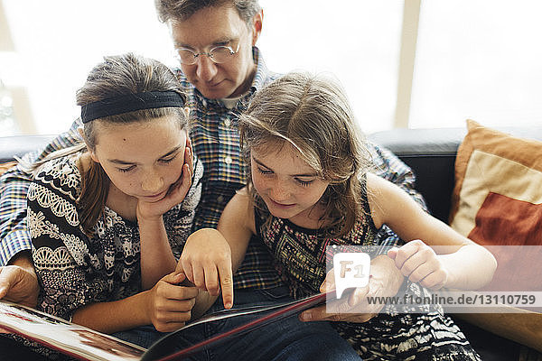 Father with daughters looking at book while sitting on sofa at home