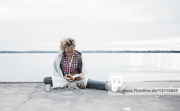 Eine Frau liest Tagebuch in voller Länge  während sie am Seeufer gegen den Himmel sitzt