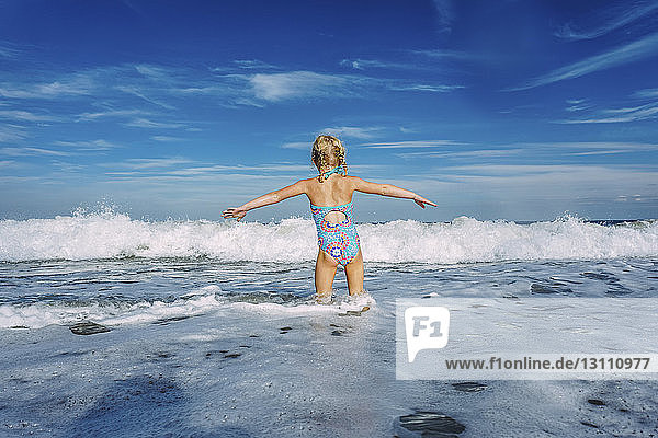 Rear view of girl with arms outstretched standing in sea against cloudy sky