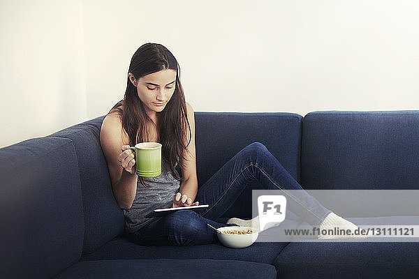 Woman using digital tablet while eating breakfast on sofa at home