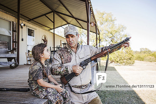 Father holding rifle and pointing while daughter sitting on porch