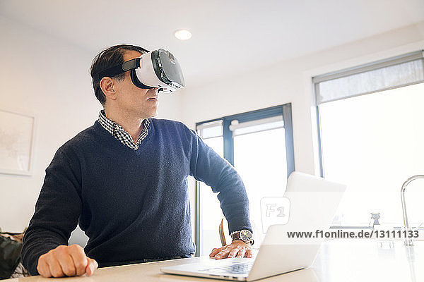 Mann mit Virtual-Reality-Simulator am Tisch sitzend mit Laptop zu Hause