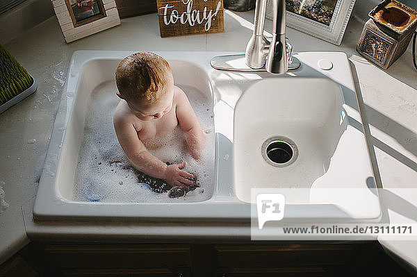 High angle view of shirtless baby boy sitting in kitchen sink at home