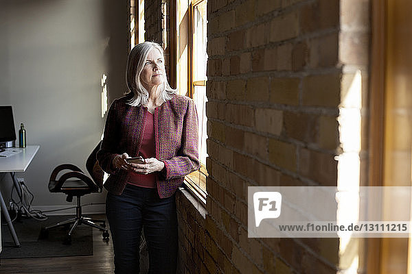 Thoughtful mature businesswoman looking through window while holding smart phone in creative office