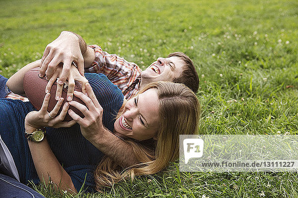 Fröhliches Paar hält Fussball in der Hand  während es auf einem Rasenfeld liegt