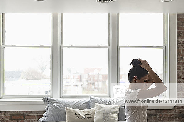 Seitenansicht einer Frau  die die Haare bindet  während sie zu Hause auf dem Bett am Fenster sitzt